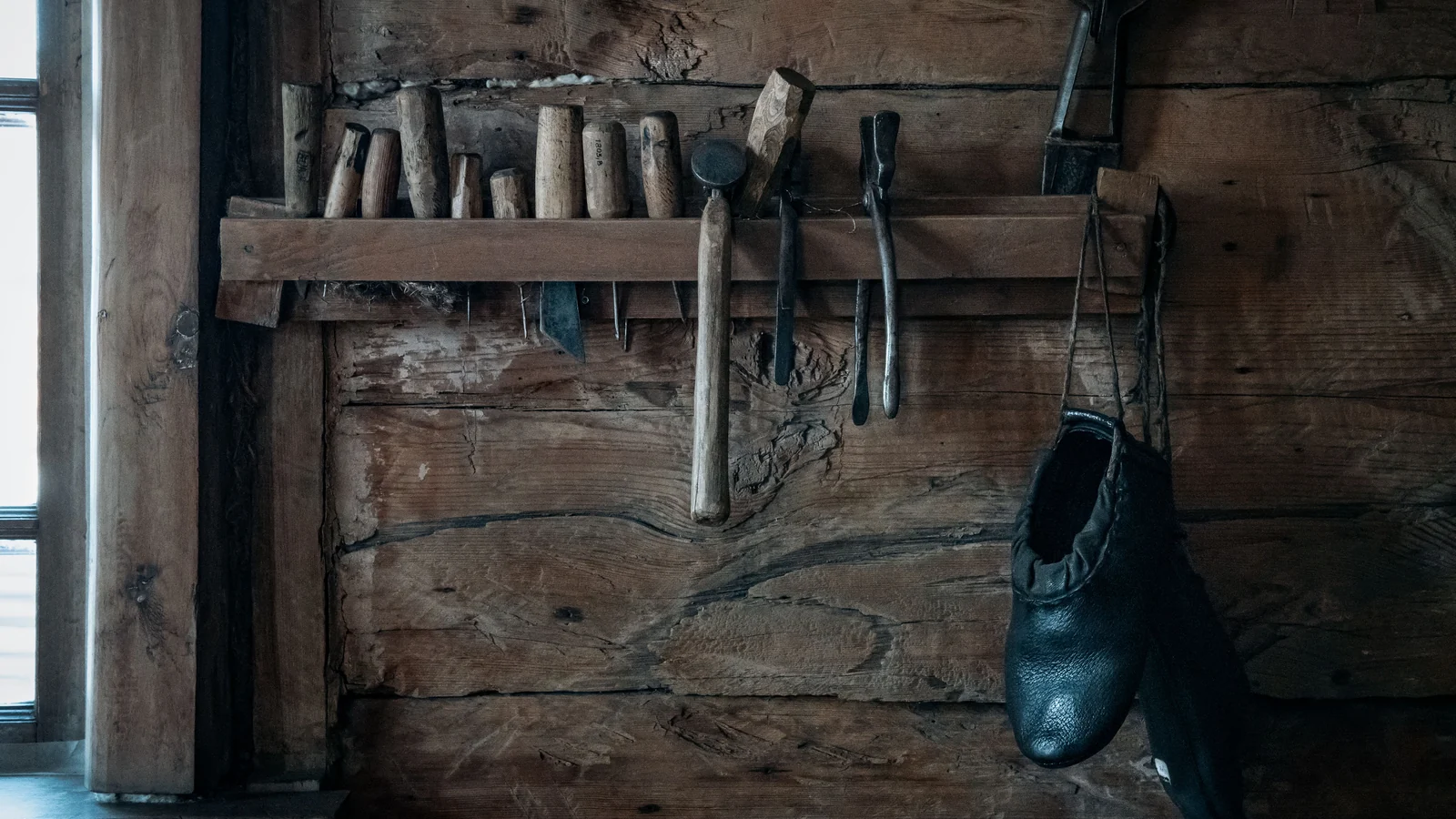 Illustrative stock image of shoemaking tools and dark shoes hanging against a weathered wooden wall.