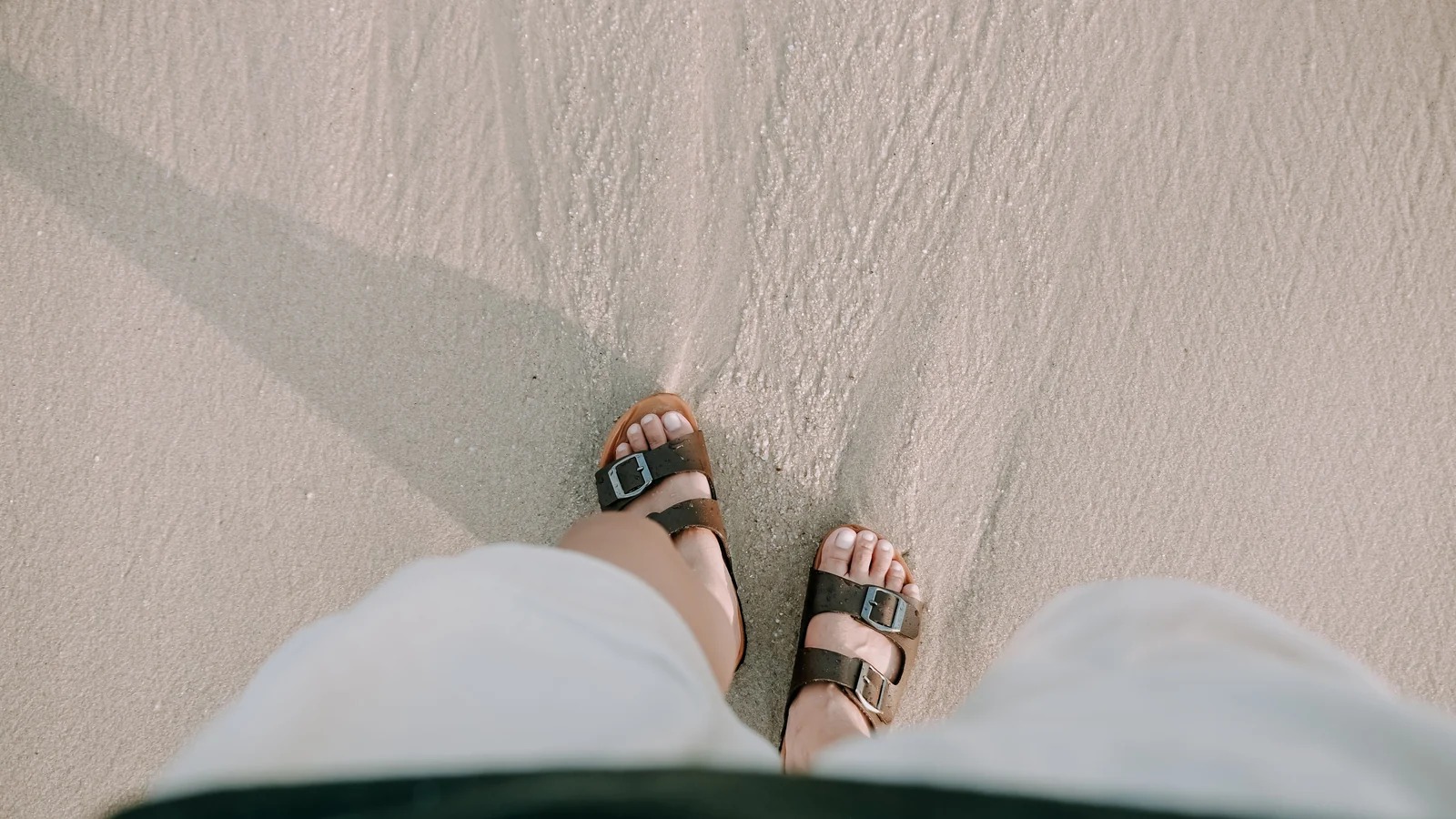Illustrative stock image looking down at feet in buckle sandals on sand.