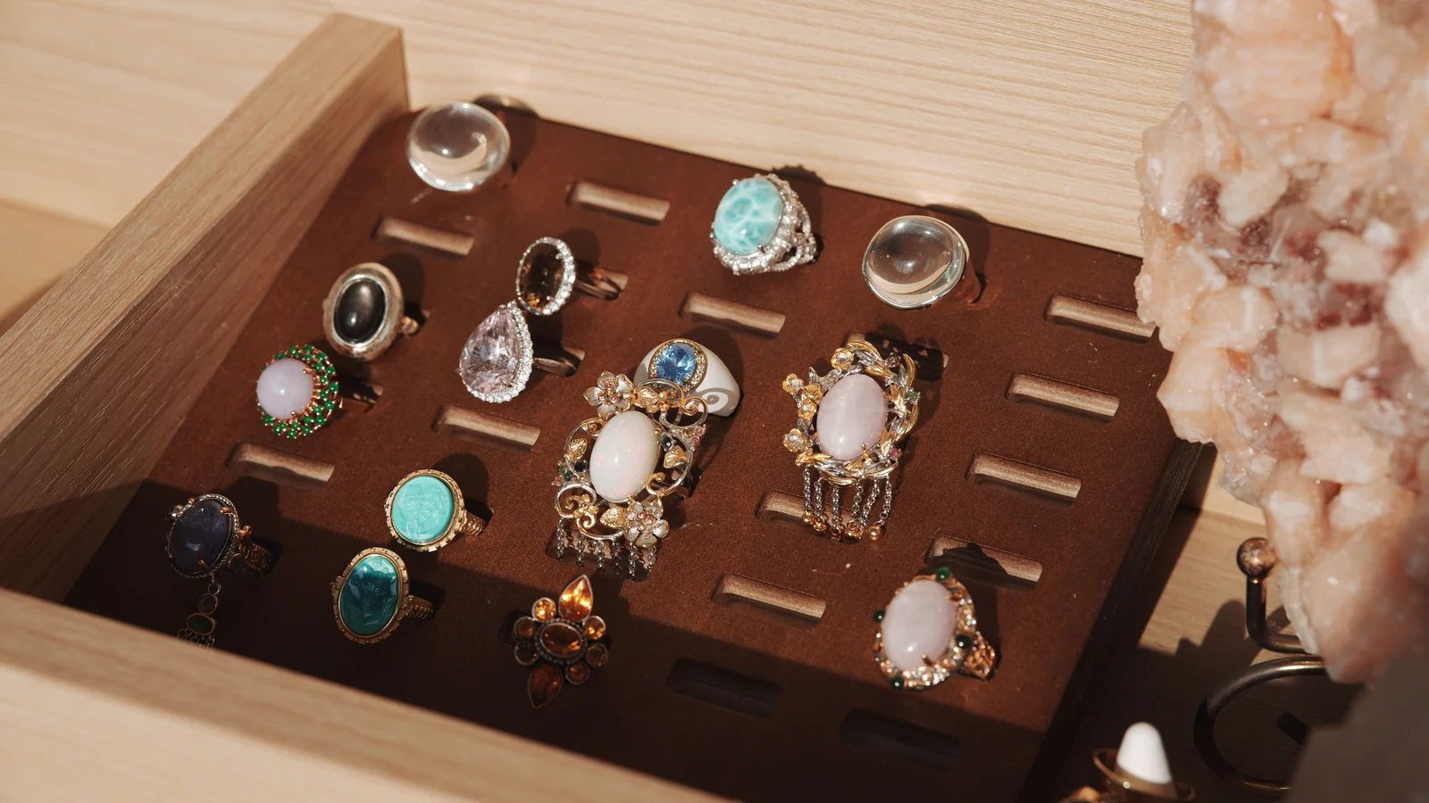 Colorful stone-set rings arranged in a wooden display tray beside a pale crystal cluster.