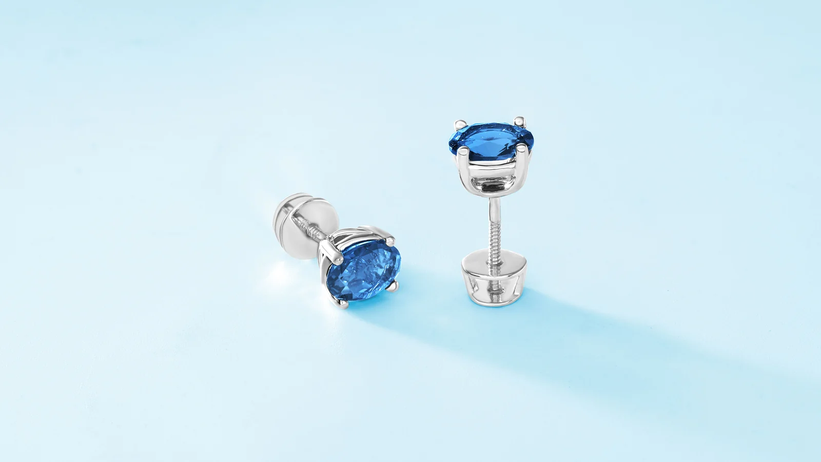 Two silver-colored stud earrings with blue stones on a pale blue surface.