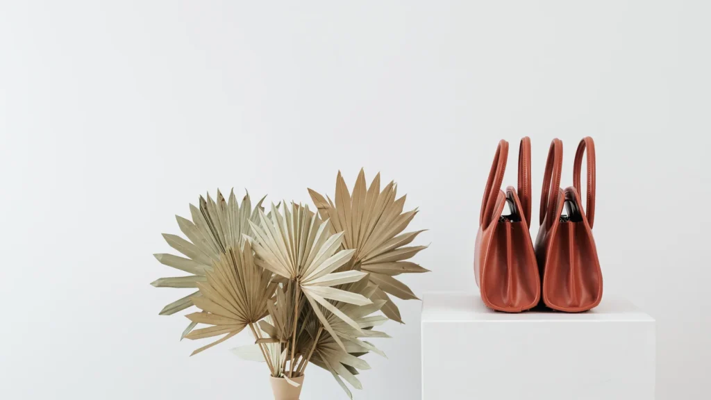Two rust-colored handbags on a white pedestal beside dried fan-shaped leaves.