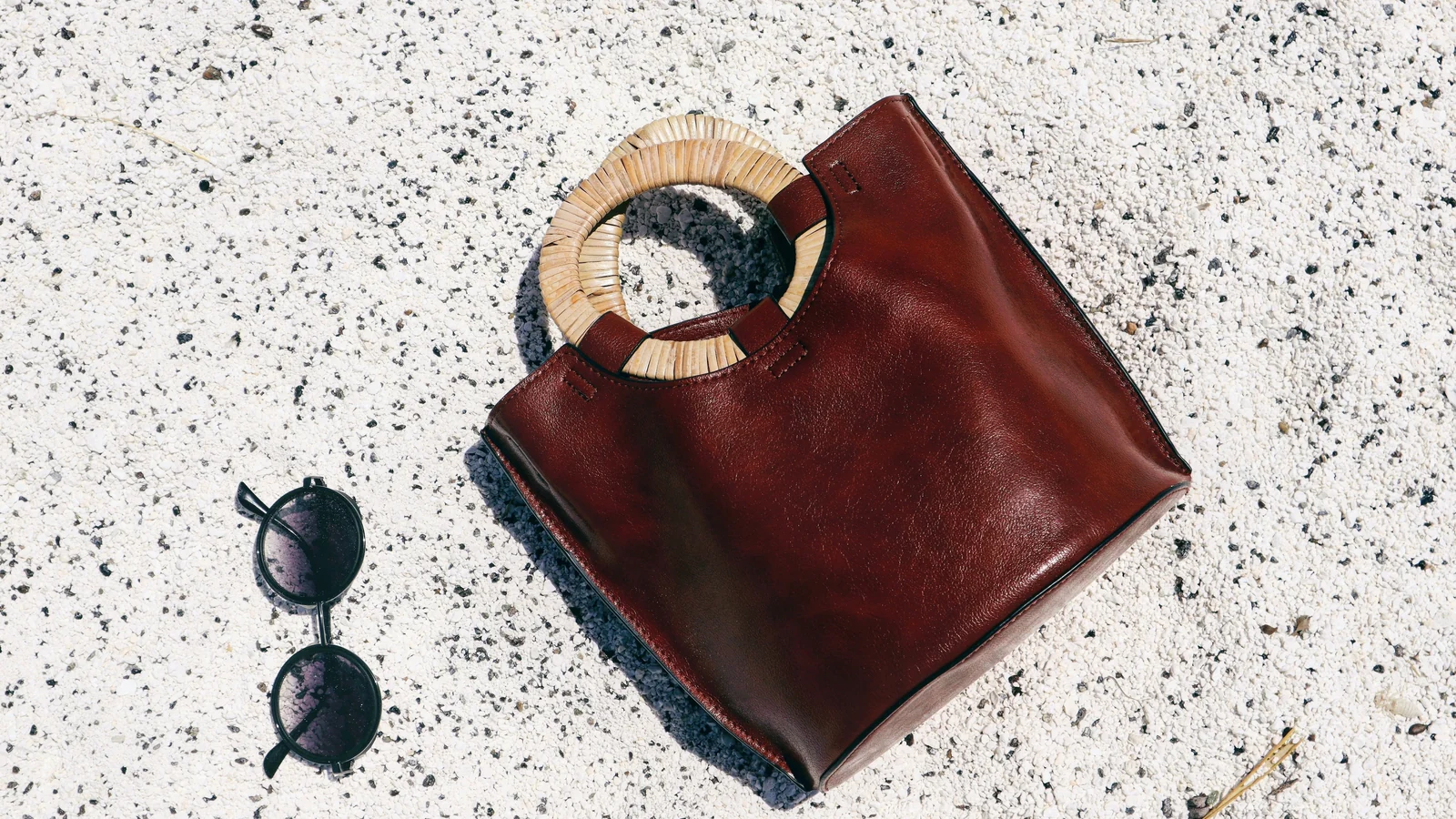 A brown handbag with round woven handles beside dark sunglasses on a textured white surface.