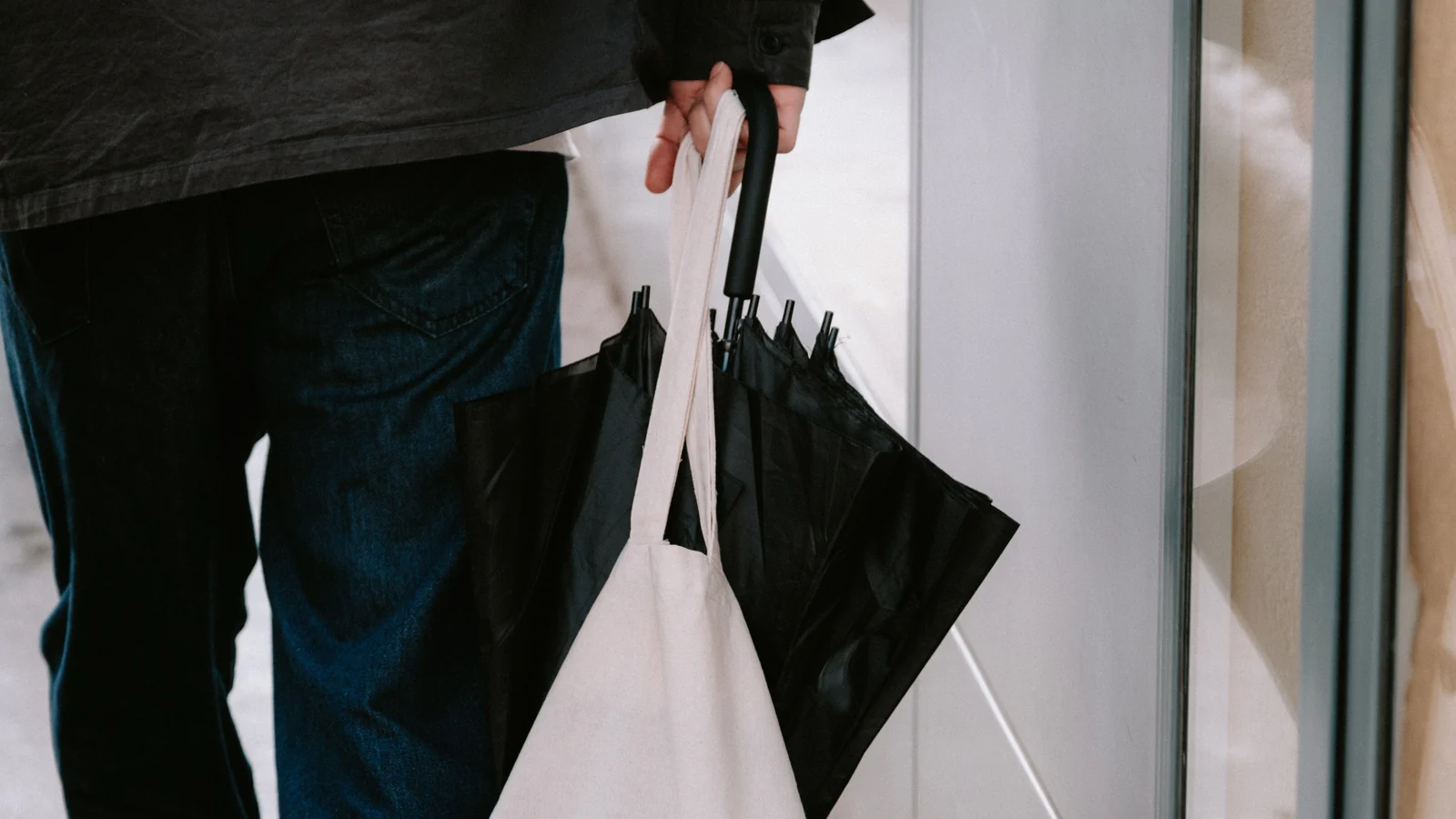 A person shown from the torso down holding a light-colored tote bag and a closed black umbrella.