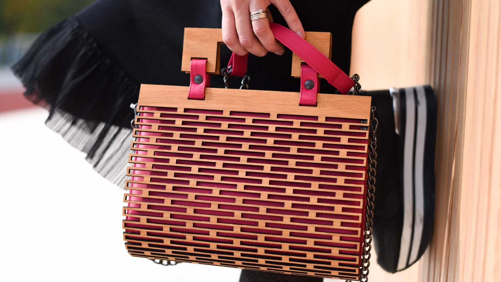 A hand holding a rectangular handbag with a wooden lattice exterior, pink straps, and a chain.